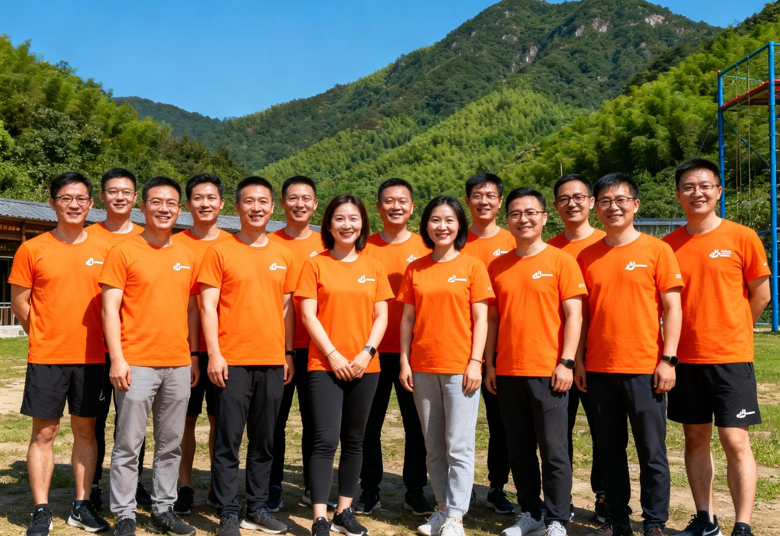 必一运动创始团队及早期核心成员在青山环抱的户外拓展基地合影，所有人穿着统一的橙色品牌T恤，面带笑容站成两排，背景是湛蓝天空和翠绿山峦，阳光温暖明亮。